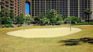 Een zandvolleyballveld met een net, omringd door gras en bomen, met een hotelgebouw op de achtergrond.