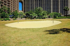 A sandy volleyball court with a net, surrounded by grass and trees, with a hotel building in the background. - Ocean Creek Unit 2424 (Myrtle Beach)