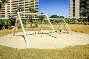 A swing set in a grassy area with a building in the background. - Ocean Creek Unit 2424 (Myrtle Beach)
