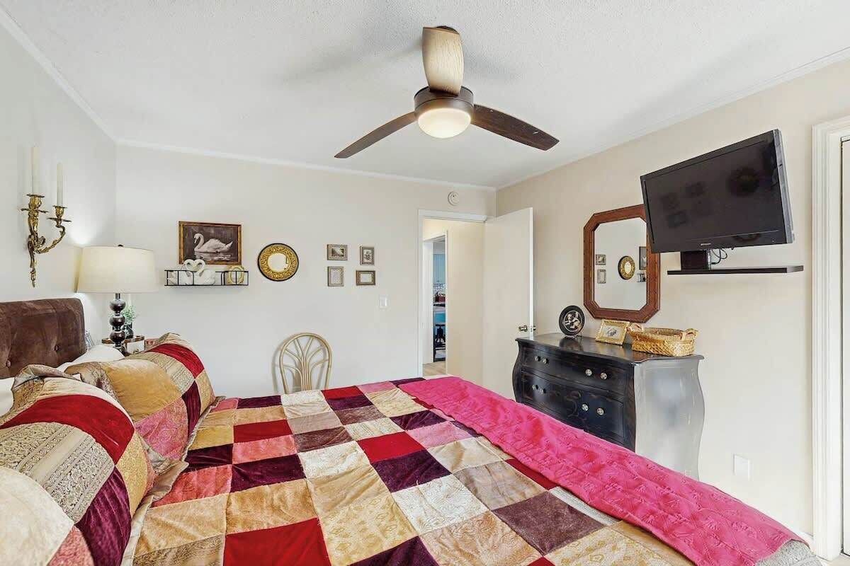 Um quarto com cama, ventilador de teto, cômoda, televisão e uma porta que dá para outro espaço.