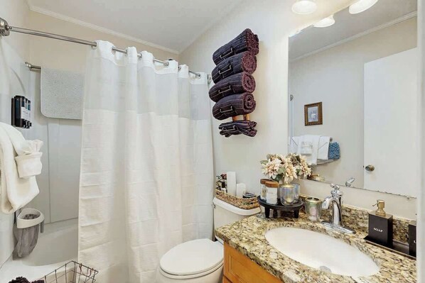A bathroom with a white toilet, a sink with a granite countertop, a shower curtain, and towels stacked on a rack. - Cozy Captains Retreat (North Myrtle Beach)