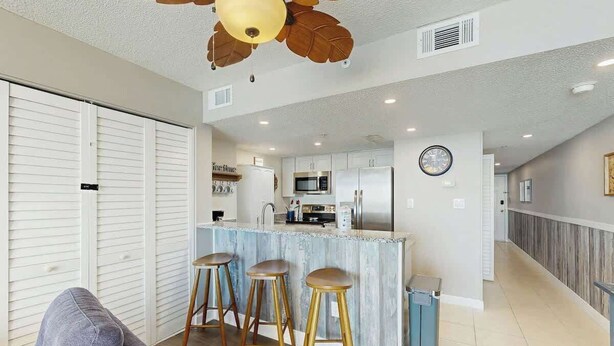 A modern kitchen with a central island, bar stools, and a ceiling fan with leaf designs. - Verandas Unit 803 Condo (North Myrtle Beach)