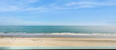 Une vue sur la plage depuis un balcon doté d’une balustrade, avec une plage de sable et l’océan en toile de fond.