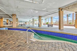 Ein überdachter Innenbereich mit einem Schwimmbecken, einem Fliesenrand, Steinböden und Tischen mit Stühlen.