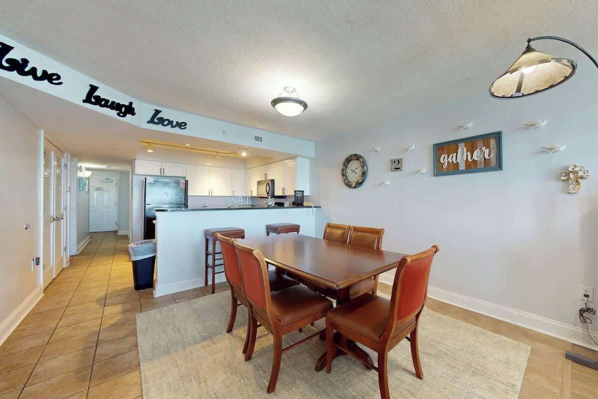 Comedor con mesa y sillas de madera, cocina al fondo y un cartel que dice 'Give Laugh Love'.