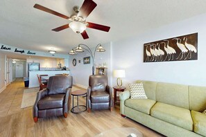 Ein Wohnzimmer mit Deckenventilator, einem grünen Sofa, zwei Ledersesseln, einem Couchtisch und einer Wanddekoration mit Vogelmotiven.