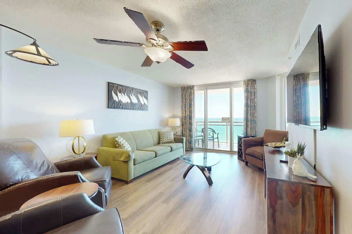 A modern living room with leather sofas, a glass coffee table, a ceiling fan, and a view to the outside.