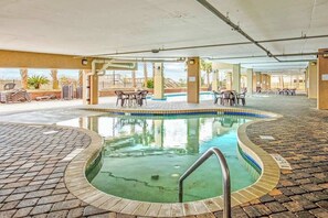 Ein Hallenbad mit gefliestem Sonnendeck, umgeben von Liegestühlen und Tischen, mit Blick auf Palmen und ein Gebäude im Hintergrund.