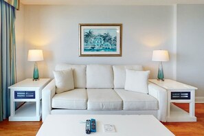 Una camera d'albergo con un divano bianco, due comodini con lampade, un quadro appeso al muro e un tavolino da caffè con telecomandi.