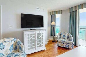 Ein Zimmer mit Fernseher, einem weißen Schrank, zwei gemusterten Sessel, einer Lampe und Meerblick durch Vorhänge.