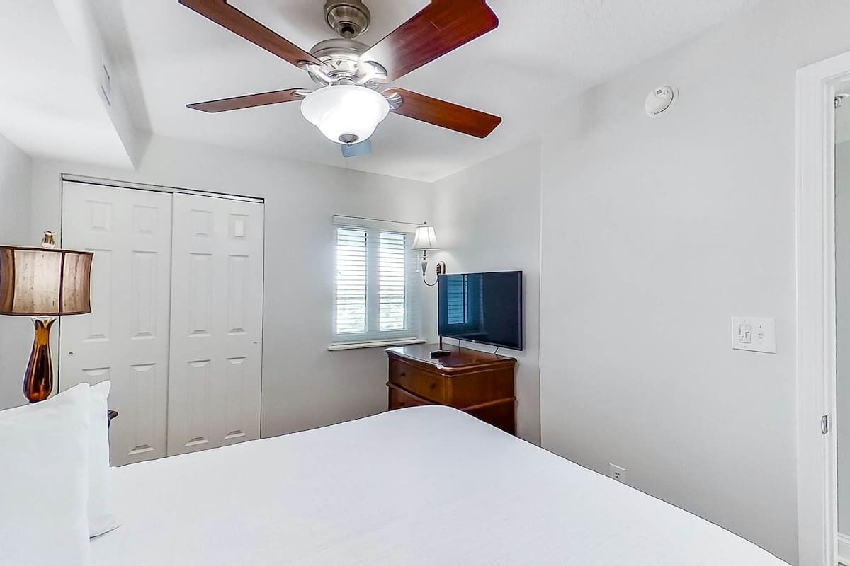 Um quarto com cama, ventilador de teto, televisão, janela com persianas e guarda-roupa.