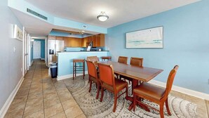 Uma sala de jantar com mesa e cadeiras, uma cozinha ao fundo e um corredor que leva a outro quarto.