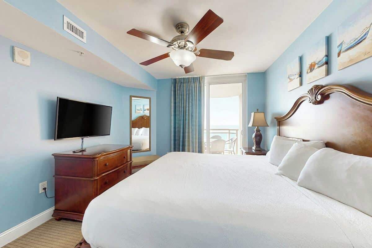 A hotel room with a large bed, a ceiling fan, a flat-screen TV, a dresser, and a window with curtains.