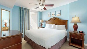 Quarto de hotel com cama, ventilador de teto, mesa de cabeceira com abajur, cômoda e janela com cortinas.