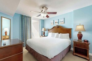 Ein Hotelzimmer mit einem Bett, einem Deckenventilator, einem Nachttisch mit Lampe, einem Schminktisch und einem Fenster mit Vorhängen.