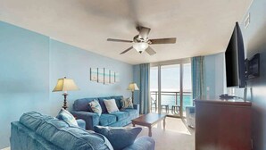 Quarto de hotel com sofá azul, televisão de tela plana, ventilador de teto e vista para o mar através de grandes janelas.