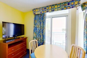 A room with a television on a wooden stand, a dining table with chairs, and a balcony with a view. - Bay Watch Resort 1410 (North Myrtle Beach)