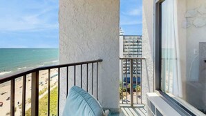Varanda com vista para a praia e oceano, uma parede branca e uma janela com cortinas.