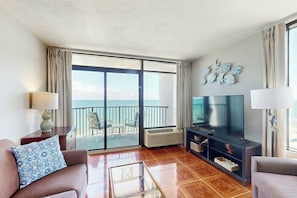 A hotel room with a balcony, a flat-screen TV, a sofa, a coffee table, and a TV stand with decorative items. - Ocean Park Resort 1222/1224 Combo (Myrtle Beach)