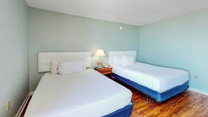 Quarto de hotel com duas camas, uma mesa de cabeceira com abajur e piso de madeira.