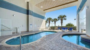 Una piscina interior con dos zonas diferenciadas, una cubierta con techo y vistas a palmeras al exterior.