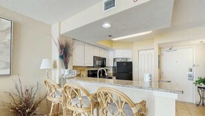 Una cocina moderna con armarios blancos, una isla central con taburetes y un comedor con un jarrón de plantas secas.