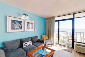 Ein Wohnzimmer mit einer Couch, einem Couchtisch und einem Sessel mit Kissen. An der Wand hängen zwei gerahmte Bilder und es gibt einen Balkon mit Blick.