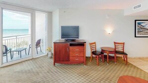 A room with a television on a wooden cabinet, a dining table with chairs, a balcony with chairs, and a wall-mounted light fixture. - Sandy Beach Resort 504 (Myrtle Beach)