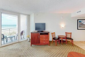 A room with a television on a wooden cabinet, a dining table with chairs, a balcony with chairs, and a wall-mounted light fixture. - Sandy Beach Resort 504 (Myrtle Beach)