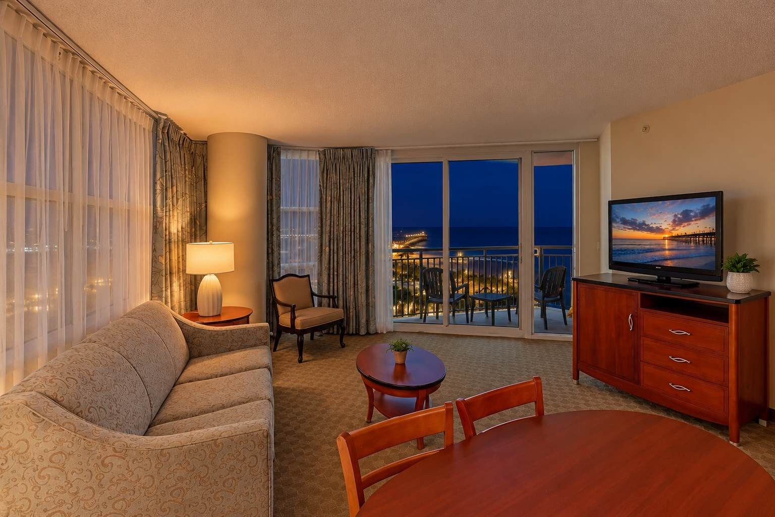 A hotel room with a sofa, a dining table, a TV, and a balcony with a view of the city lights.