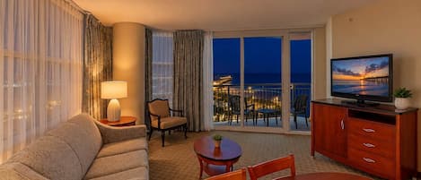 A hotel room with a sofa, a dining table, a TV, and a balcony with a view of the city lights.