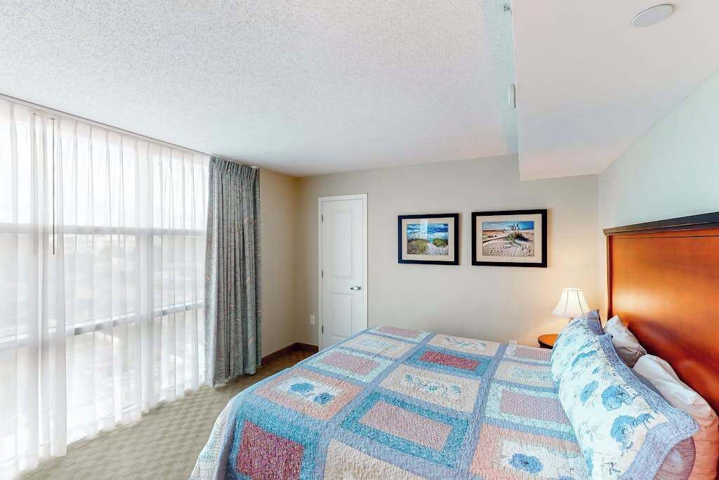 A hotel room with a bed, a window with curtains, a door, and two framed pictures on the wall.