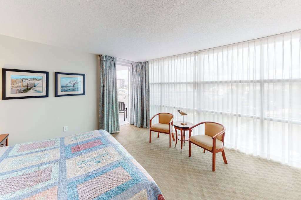 Ein Hotelzimmer mit einem großen Bett, zwei Stühlen, einem kleinen Tisch und Blick durch leichte Vorhänge.