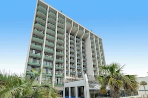 Ein mehrstöckiges Hotel mit Balkonen, einem zentralen Eingang und Palmen im Vordergrund.