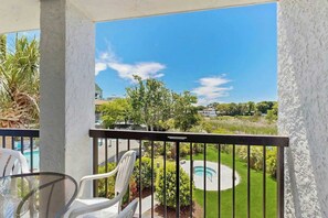 Ein Balkon mit Glastisch und Stühlen, ein Pool und Blick auf eine grüne Landschaft mit Bäumen und blauem Himmel.
