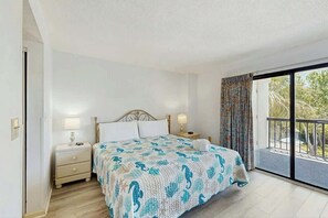A bedroom with a bed, two bedside tables, a window with curtains, and a view of the outdoors. - Sea Mark Tower Unit 205 (Myrtle Beach)