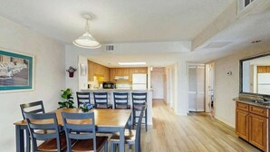 A dining area with a wooden table and chairs, a kitchen in the background, and a hallway leading to another room. - Sea Mark Tower Unit 205 (Myrtle Beach)