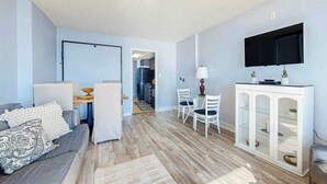 A modern living room with a grey sofa, a dining area with white chairs, a flat-screen TV mounted above a white cabinet, and a wooden floor. - Ocean Park Resort 0922/0924 Combo (Myrtle Beach)