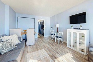 A modern living room with a grey sofa, a dining area with white chairs, a flat-screen TV mounted above a white cabinet, and a wooden floor. - Ocean Park Resort 0922/0924 Combo (Myrtle Beach)