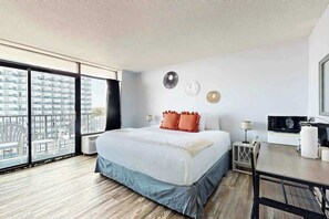 Habitación de hotel con una cama grande, un escritorio, una silla y vistas a un paisaje urbano a través de ventanas amplias.