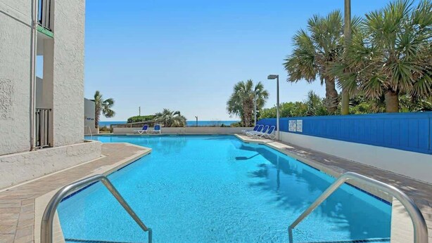 A swimming pool with clear blue water, surrounded by a blue fence, palm trees, and a view of the ocean. - Ocean Park Resort 0922/0924 Combo (Myrtle Beach)