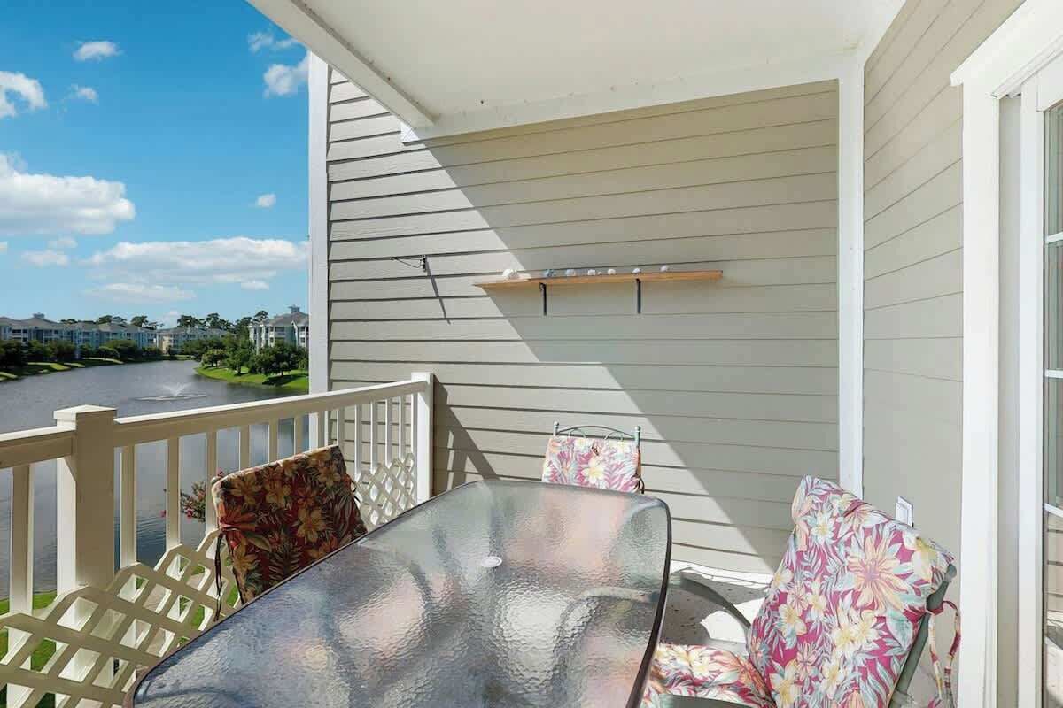 A balcony with a glass table and two chairs, a white railing, and a view of a road and greenery.