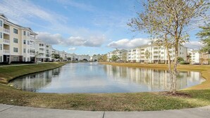 Un quartier résidentiel caractérisé par un plan d’eau paisible où se reflètent les bâtiments, un ciel dégagé et une pelouse bien entretenue.