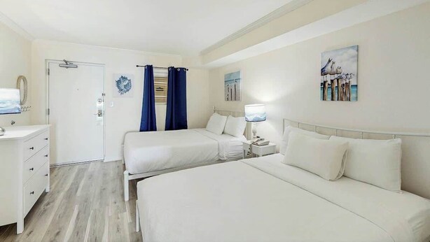 Quarto de hotel com duas camas, uma escrivaninha, uma cadeira, uma mesinha de cabeceira e uma porta.