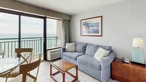 A hotel room with a balcony, a sofa, a coffee table, a chair, and a bedside table with a lamp. - Sandcastle South 1509 (Myrtle Beach)