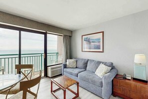 A hotel room with a balcony, a sofa, a coffee table, a chair, and a bedside table with a lamp. - Sandcastle South 1509 (Myrtle Beach)