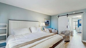 Un dormitorio con una cama grande, una mesita de noche, una silla y una puerta que da a otra habitación.