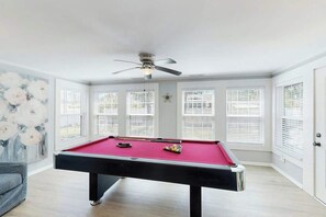 A pool table with a ceiling fan, large windows with blinds, and a painting on the wall. - Coastal Retreat on Bay Street (North Myrtle Beach)