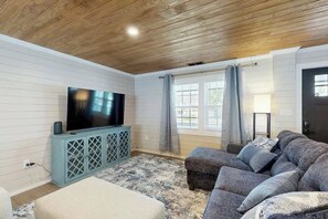 A living room with a television on a cabinet, a sofa, and a window with curtains. - Coastal Retreat on Bay Street (North Myrtle Beach)