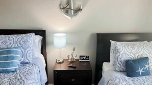 A hotel room with two beds, each with patterned bedding, a nightstand with a lamp, an anchor decoration, and a clock. - River Oaks Unit 25A Condo (Myrtle Beach)
